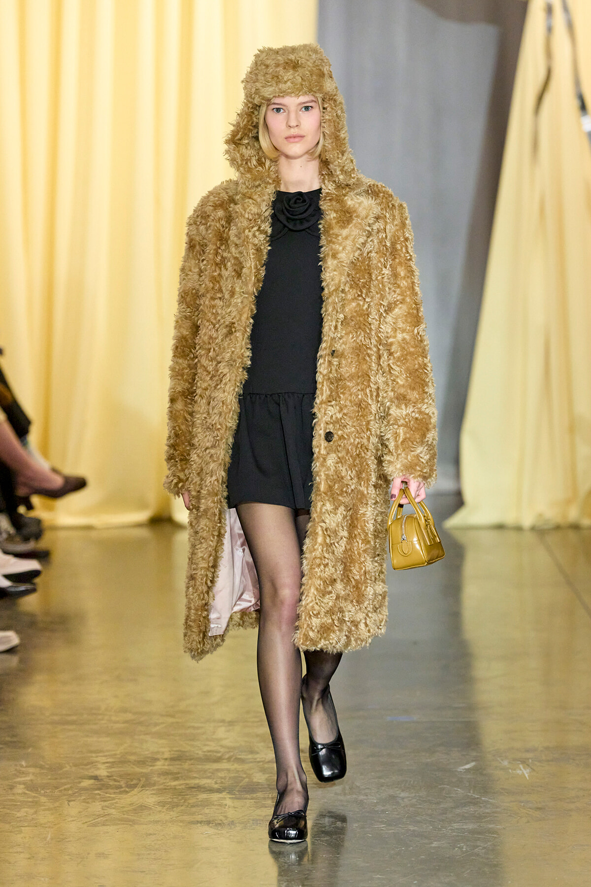 Model walking runway in a long tan fuzzy coat over a black dress, carrying a small tan handbag.