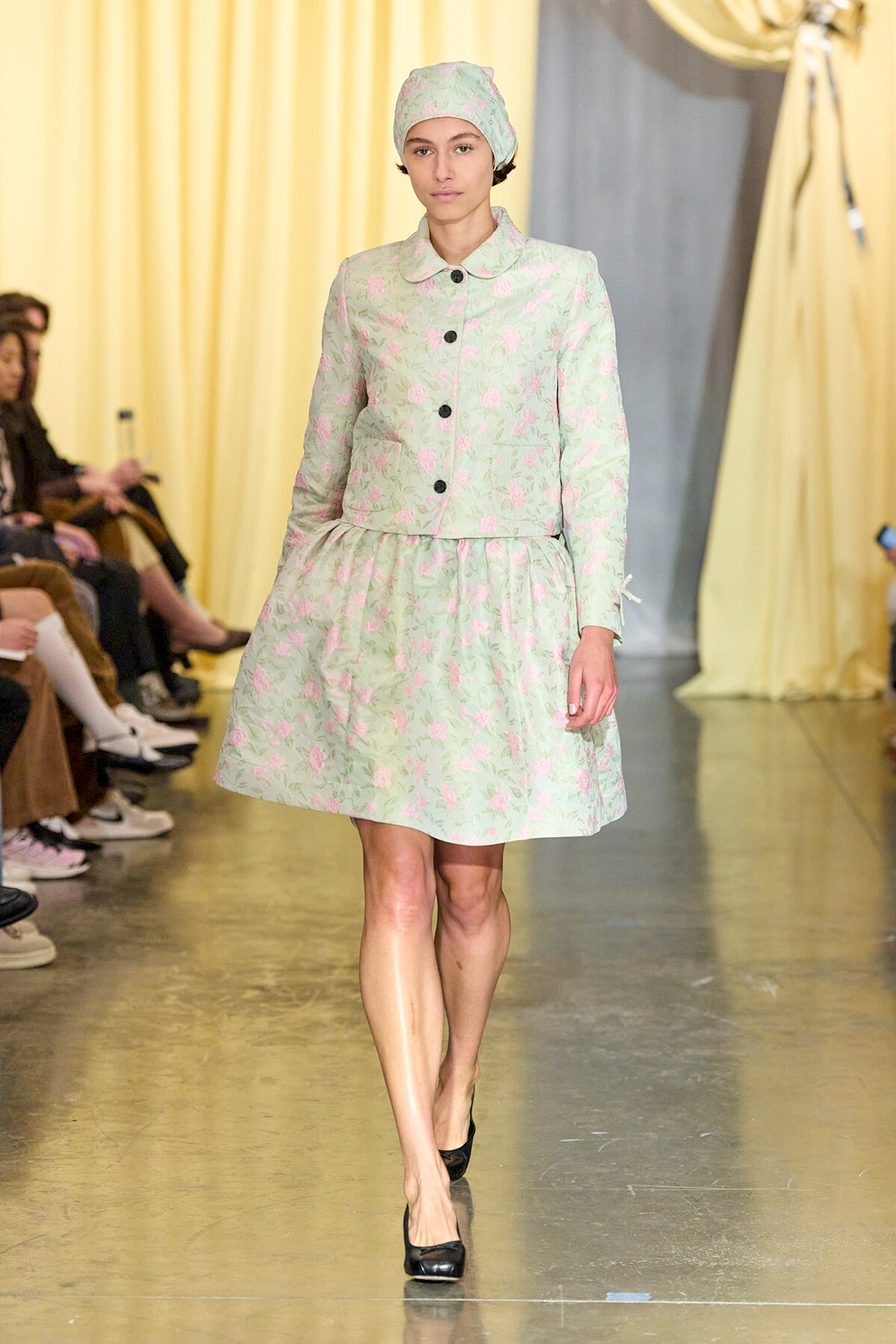 Model walks the runway in a mint green floral coat-dress with a matching headscarf and black buttons on the front.