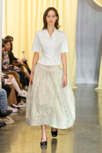 Model walking runway in a white short-sleeve blazer and mint floral ankle-length skirt; audience on the left.