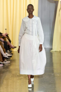 Model walks a fashion runway in a white pinstripe blouse with a large white collar and a full white skirt, wearing white shoes.