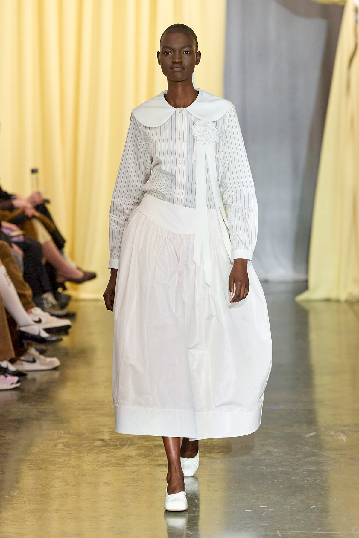 Model walks a fashion runway in a white pinstripe blouse with a large white collar and a full white skirt, wearing white shoes.