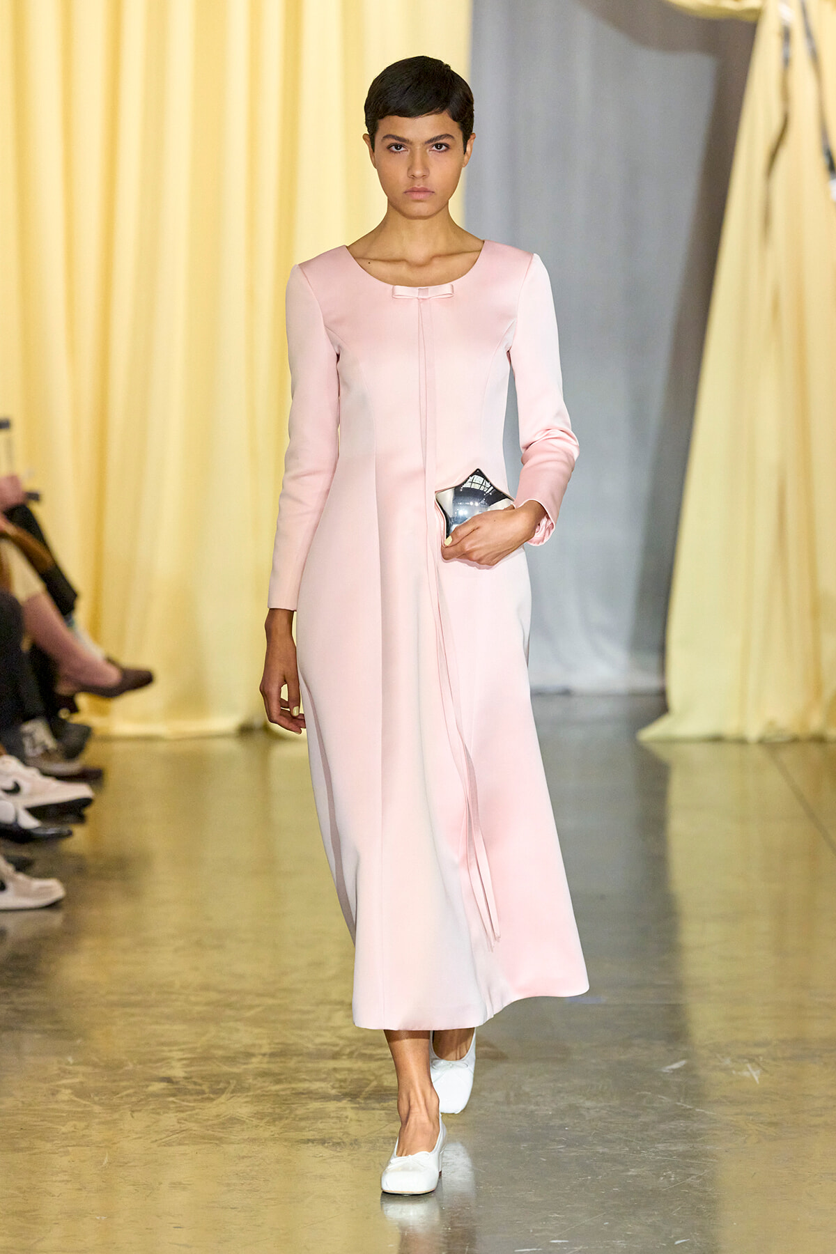 Model walking a runway in a pale pink long-sleeve midi dress, holding a small metallic clutch and wearing white shoes.