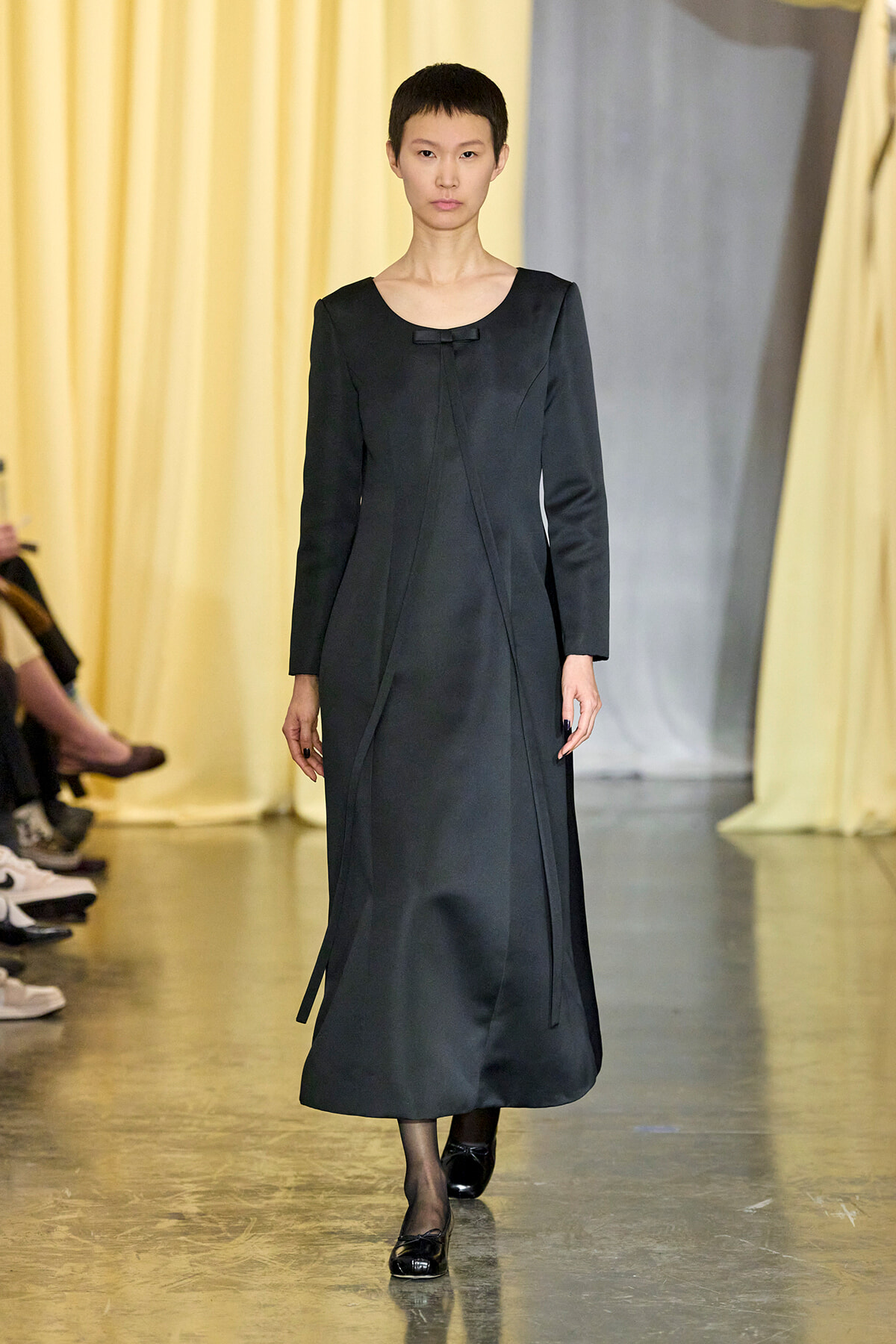 Model walking a fashion runway in a long black dress with a small bow at the neckline.
