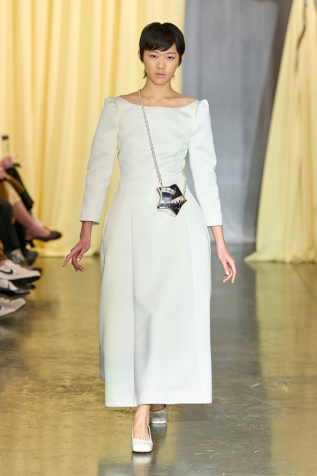 Model walks the runway wearing a pale blue long dress with a square neckline and puff sleeves, carrying a star-shaped metallic bag on a chain.