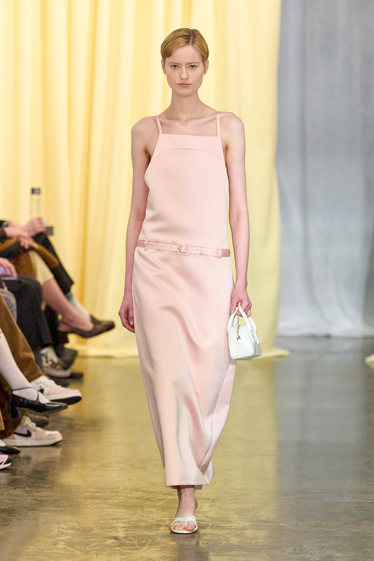 Model walking a fashion runway in a pale pink sleeveless midi dress with a thin belt, carrying a small white handbag and wearing white sandals.