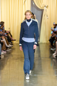 Female model walking a fashion show runway in a navy zip-up hoodie with rose appliques on the shoulder, matching navy pants, and a striped shirt peeking at the hem, white sneakers.