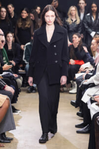 Model walks a fashion runway in a black double-breasted coat; audience sits on both sides watching.