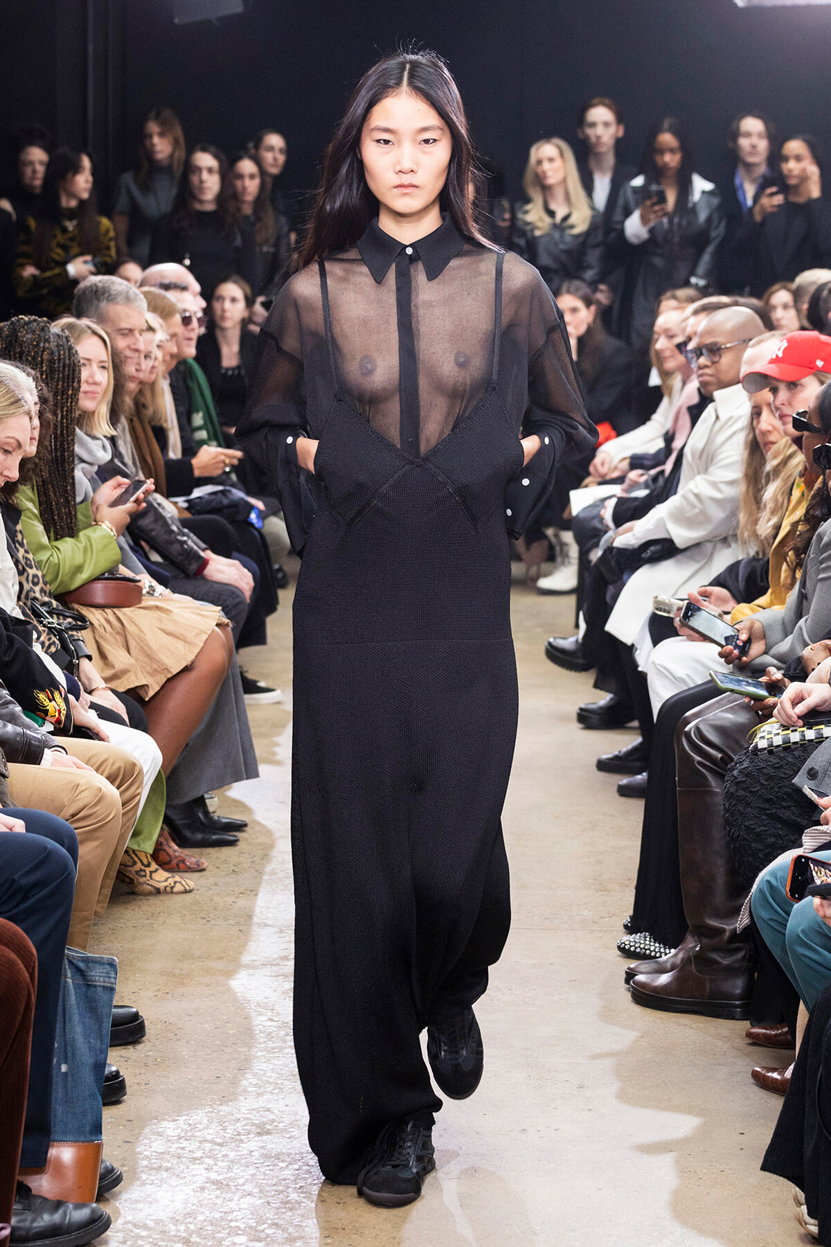 Model walks the runway in a sheer black blouse over a black dress, with a seated audience on both sides watching intently.