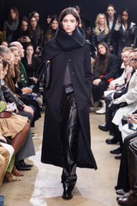 Model walks a runway in a long black coat with an oversized high-neck collar and black leather pants, as seated audience watch.