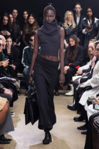 Model walks a fashion show wearing a dark gray sleeveless knit top with a large cowl neck and a long black skirt, carrying a black handbag while seated audience looks on.