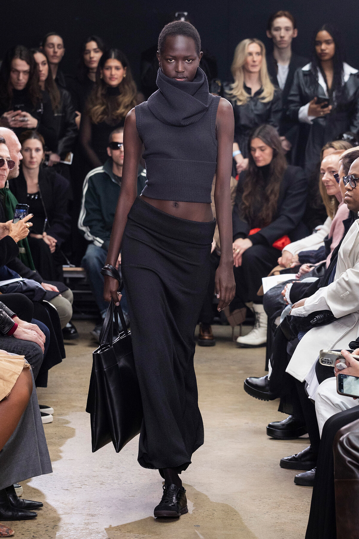 Model walks a fashion show wearing a dark gray sleeveless knit top with a large cowl neck and a long black skirt, carrying a black handbag while seated audience looks on.