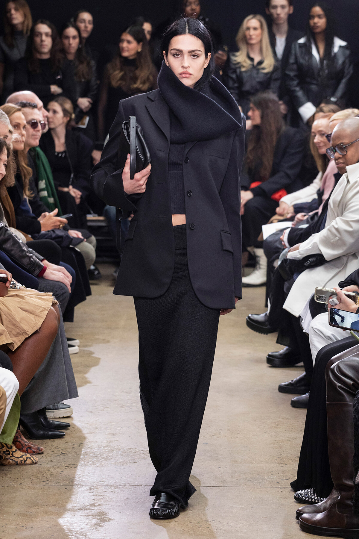 Model walks a fashion runway in an oversized black coat with a large scarf, holding a black clutch; seated audience lines the sides.