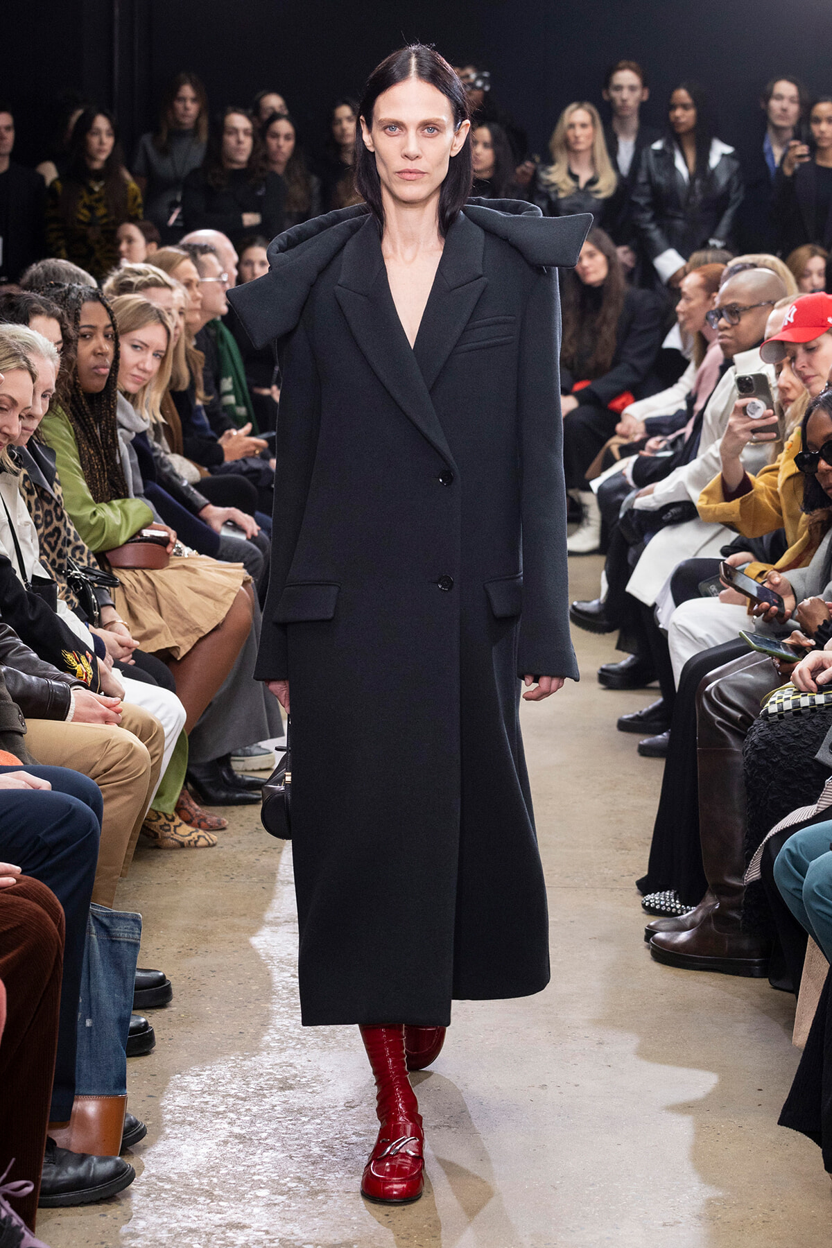 Model walks a runway wearing an oversized black coat with dramatic padded shoulders and red shin-high boots; audience seated on both sides.