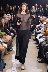 Model walks down the runway in a sheer black top and black skirt, clutch in hand, while the audience watches.