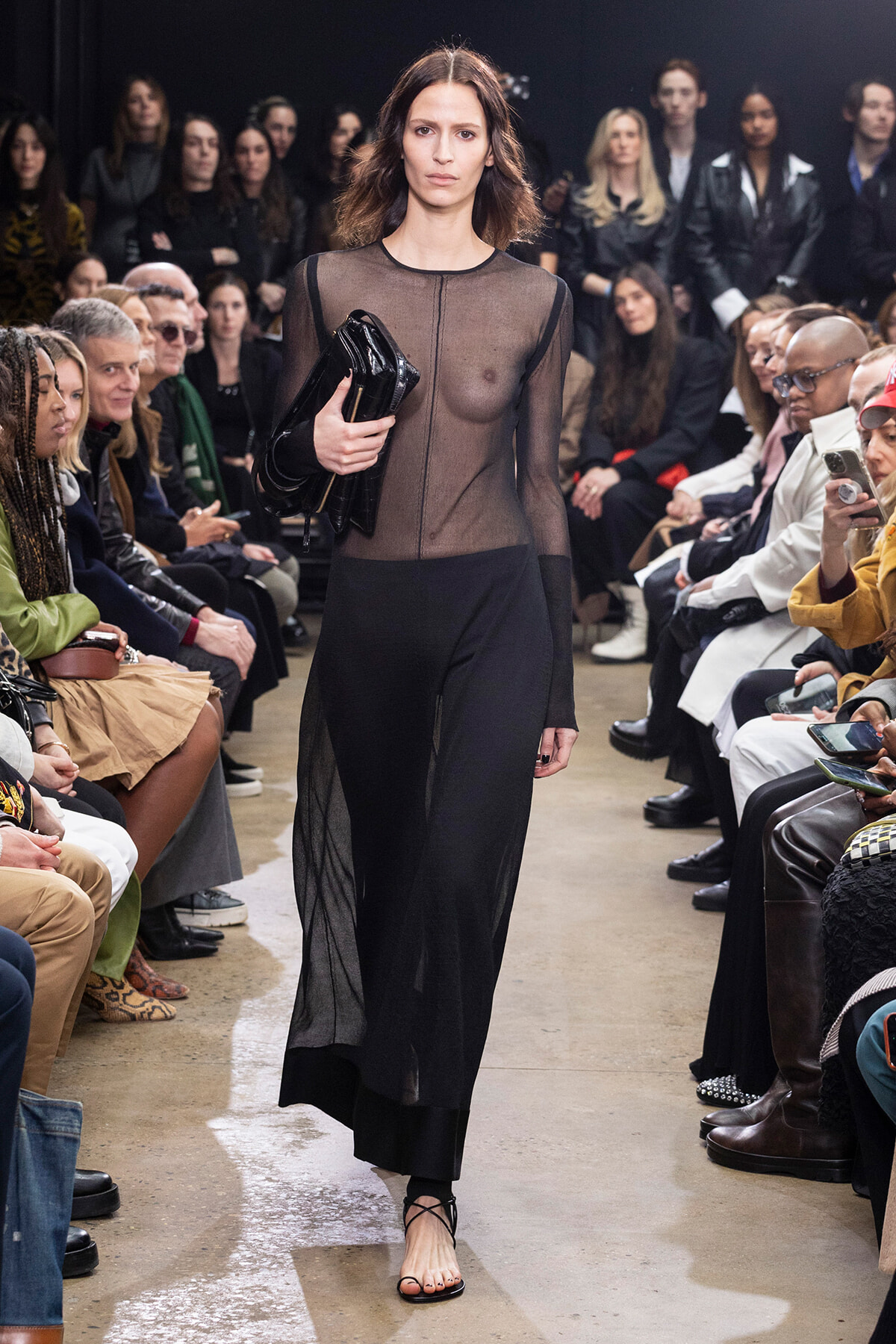 Model walks down the runway in a sheer black top and black skirt, clutch in hand, while the audience watches.