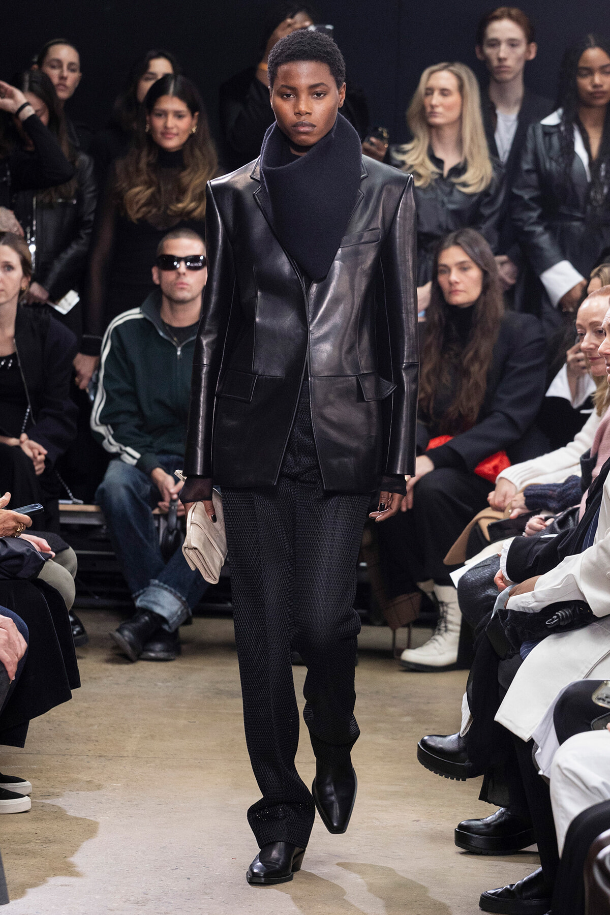 Model walking the runway in a black leather blazer and oversized scarf; seated audience behind.