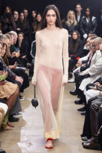 Model wearing a sheer beige long-sleeve gown walking a runway with a seated audience on both sides.