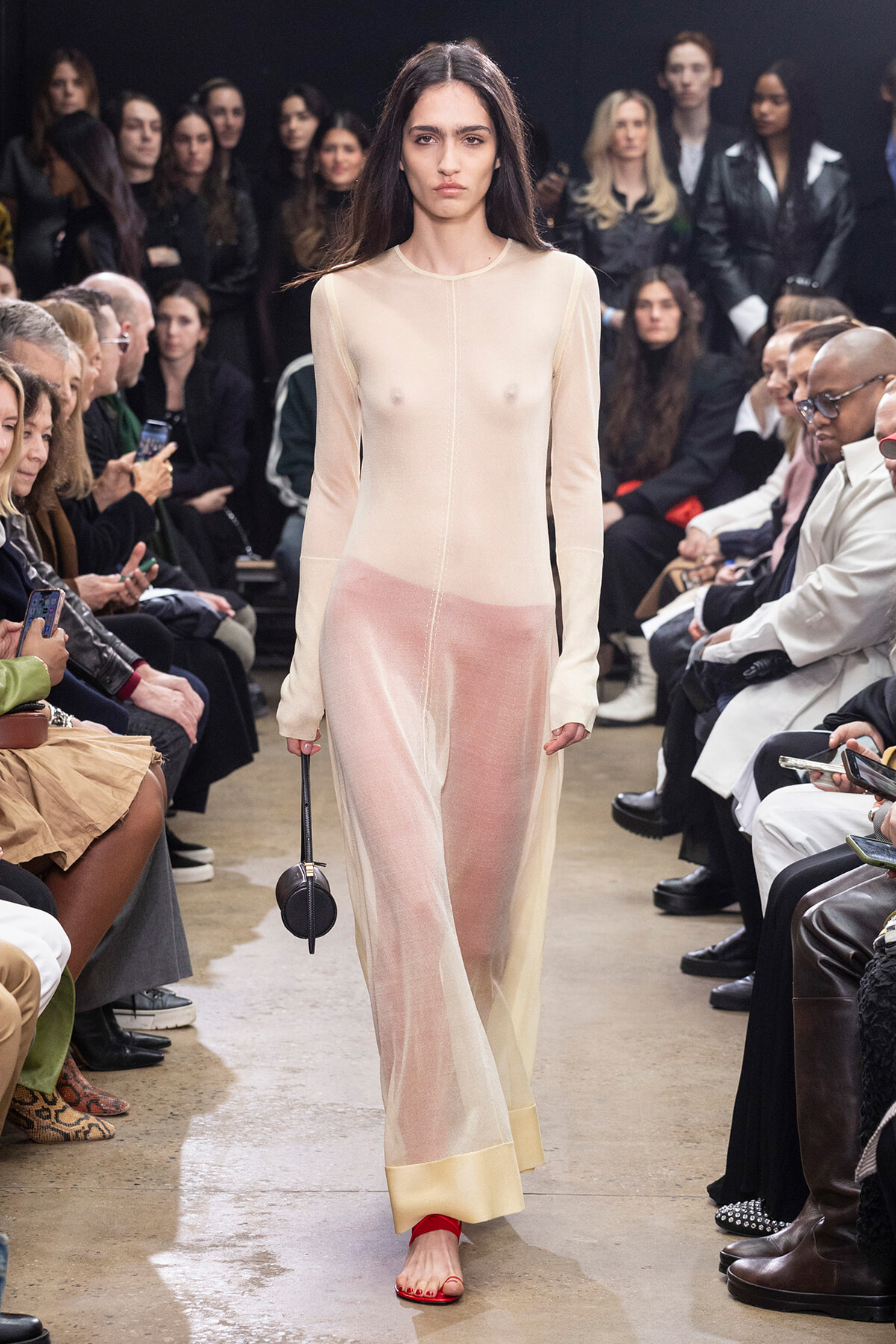 Model wearing a sheer beige long-sleeve gown walking a runway with a seated audience on both sides.