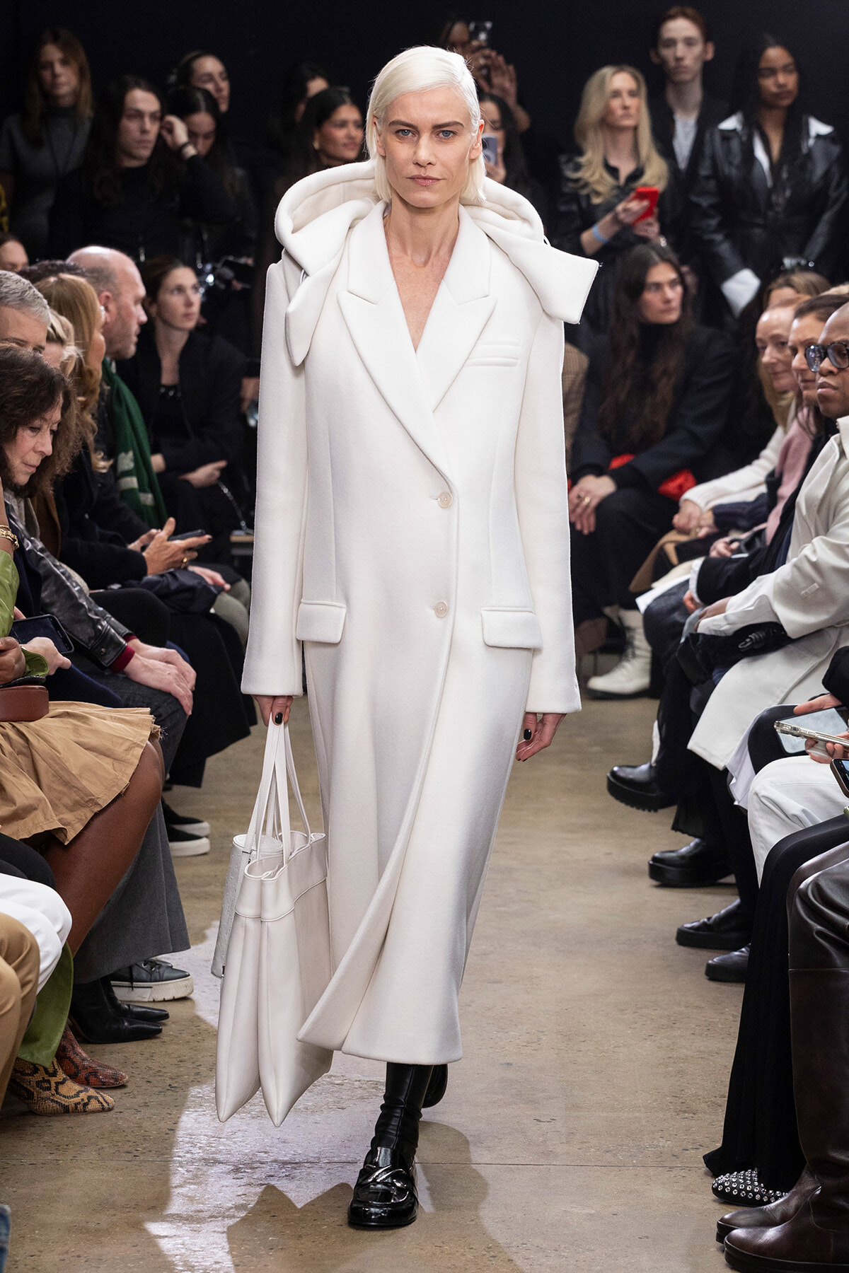 Model walks the runway in a white oversized coat with a hood, carrying a white handbag; audience seated along the sides.