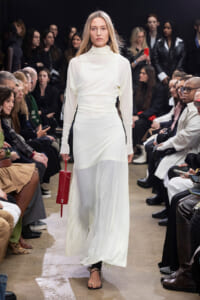 Model walks the runway in a white, high-neck gown while holding a red clutch; audience watches from both sides.