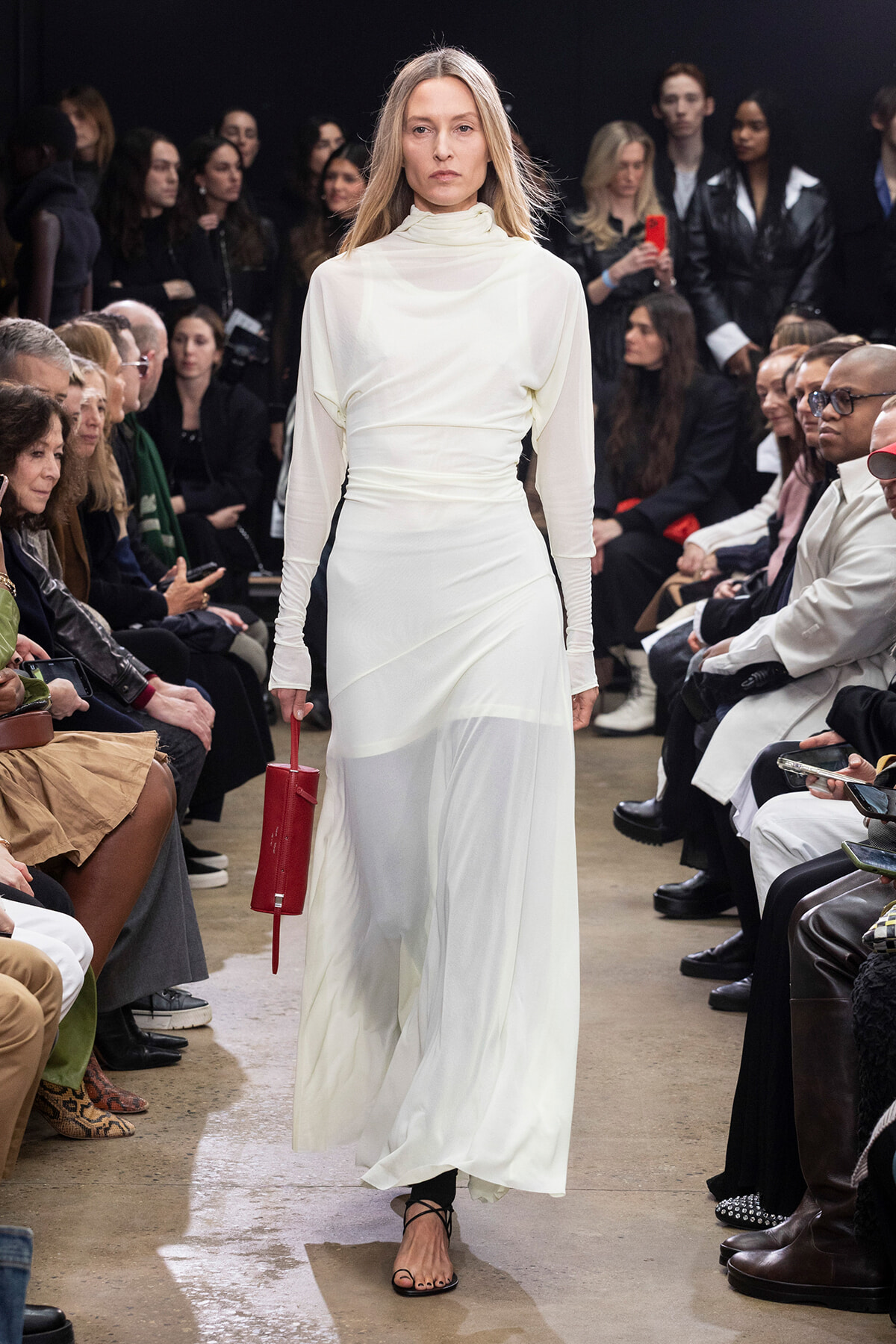 Model walks the runway in a white, high-neck gown while holding a red clutch; audience watches from both sides.