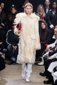 Model on runway wearing a cream fur coat and white boots, holding a red clutch as spectators watch the show.