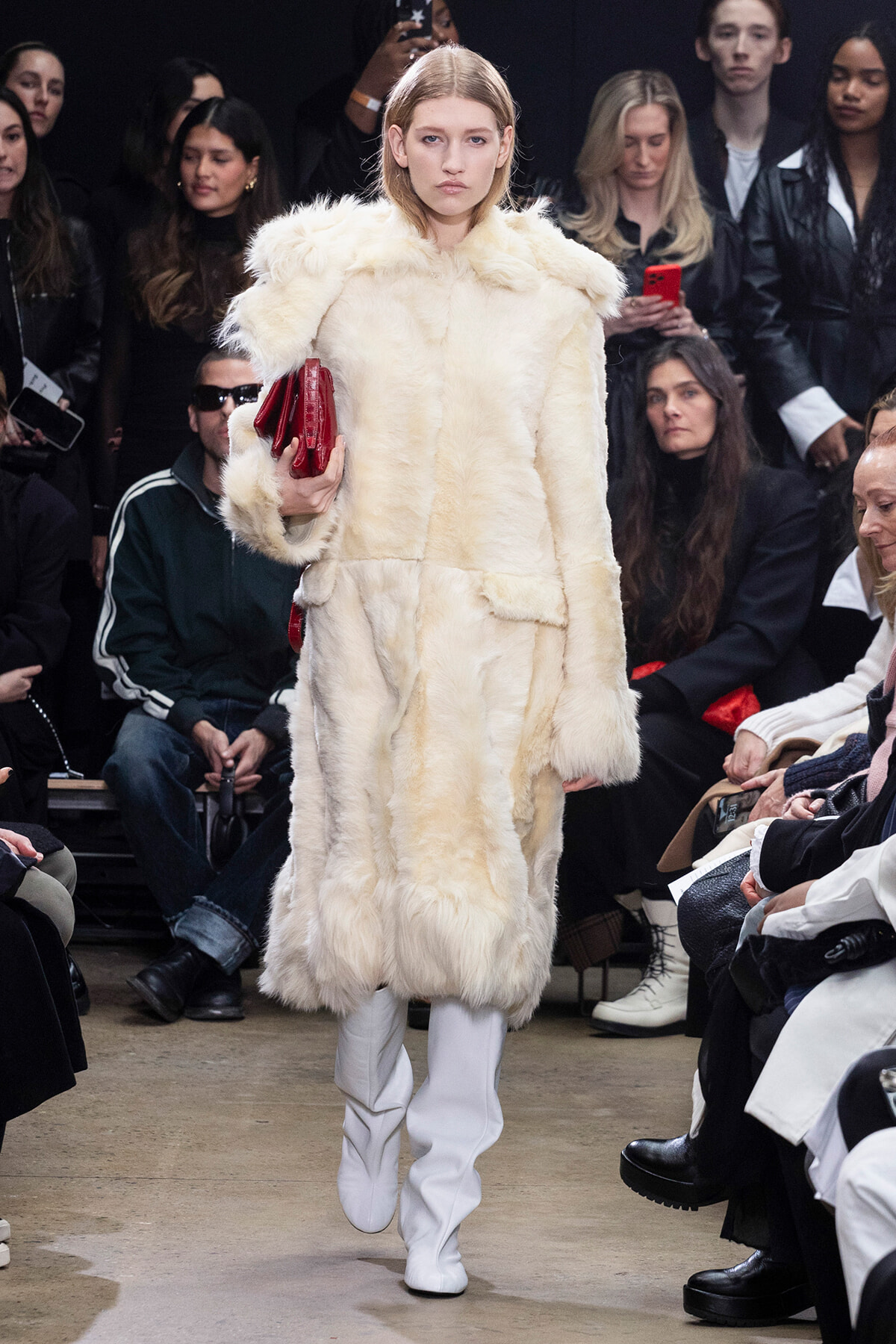 Model on runway wearing a cream fur coat and white boots, holding a red clutch as spectators watch the show.