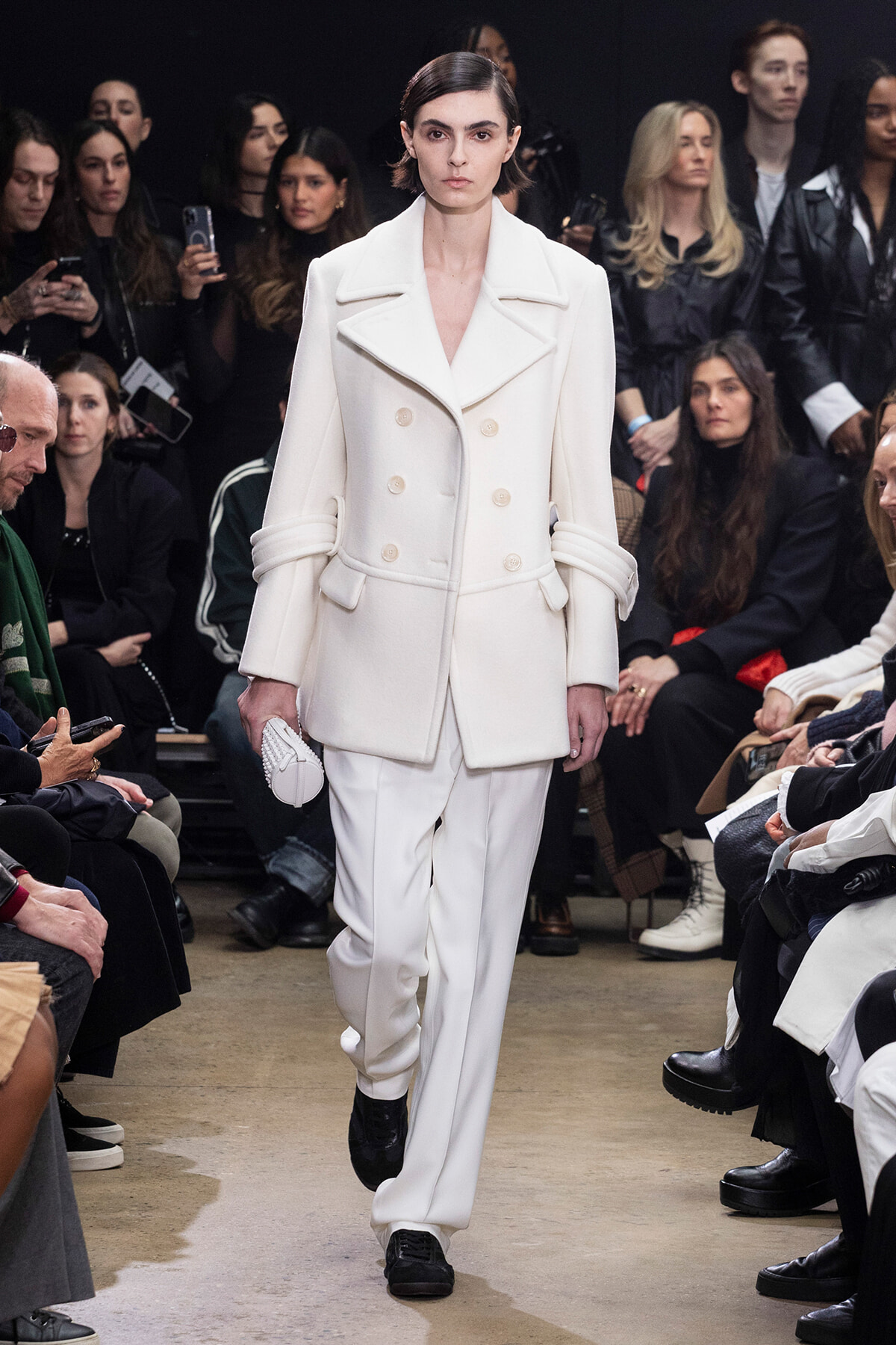 Model walks a runway in an all-white double-breasted coat and matching pants, holding a small white clutch.