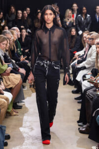Model walks a runway in a sheer black blouse, paint-splattered black jeans, red shoes, and a small metallic clutch, with an audience watching.
