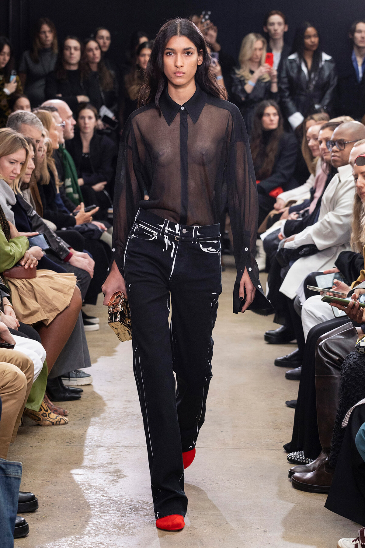 Model walks a runway in a sheer black blouse, paint-splattered black jeans, red shoes, and a small metallic clutch, with an audience watching.