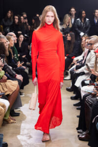 Model walks the runway in a bright red, long-sleeve turtleneck dress; audience watches from both sides.