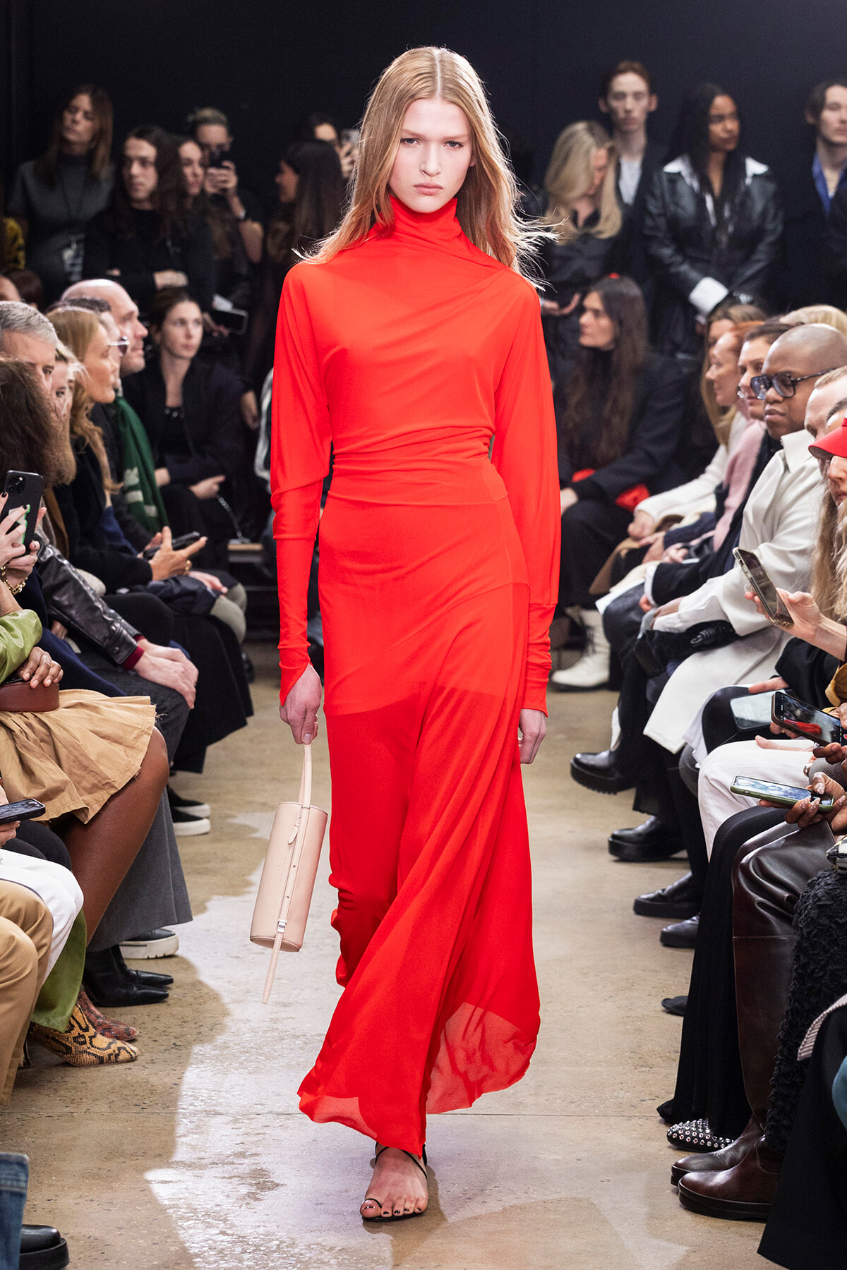 Model walks the runway in a bright red, long-sleeve turtleneck dress; audience watches from both sides.