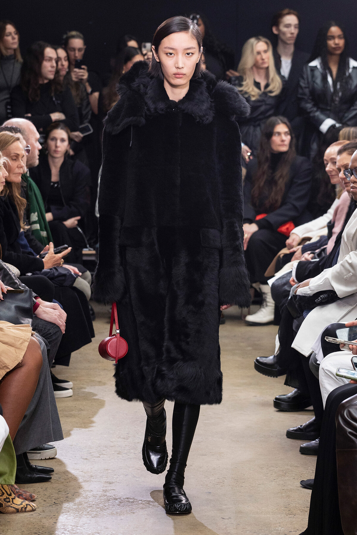 Model walks the runway in a long black fur coat with a red circular handbag, flanked by seated audience members on both sides.