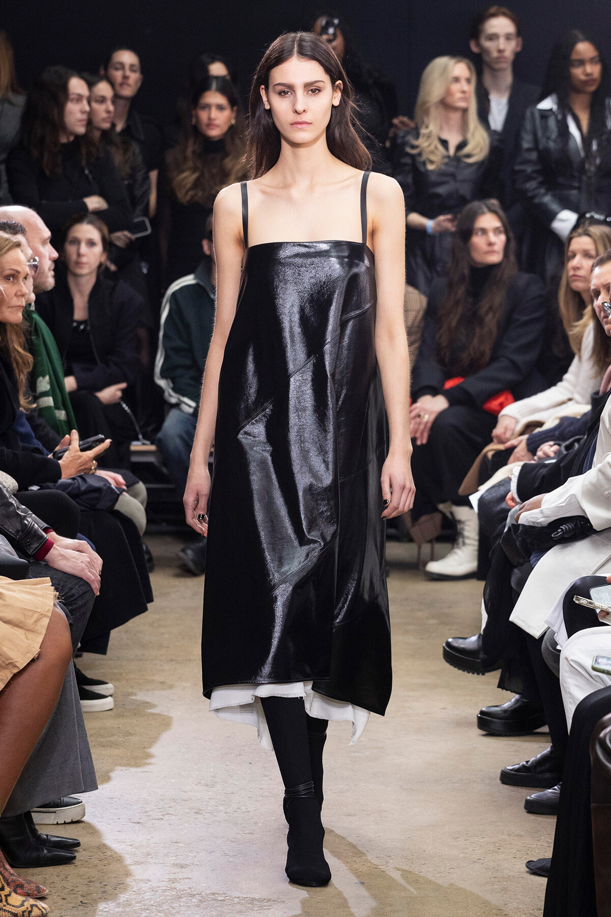 Model walking a fashion show runway in a black shiny slip dress with thin straps, audience seated on both sides.