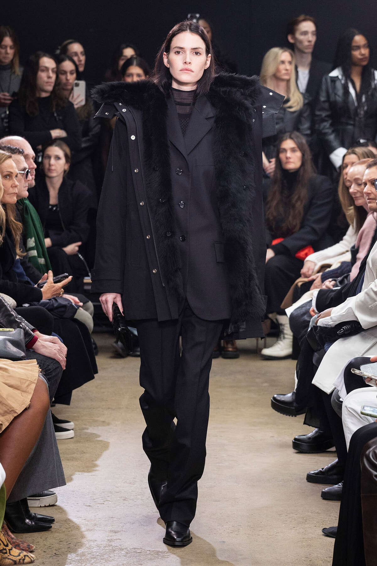 Model walking the runway in a black fur-trimmed coat; audience seated along the sides watches.