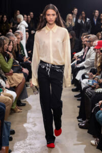Female runway model walking a fashion show in a sheer beige blouse, black paint-splattered pants, and bright red shoes, with seated audience on both sides.