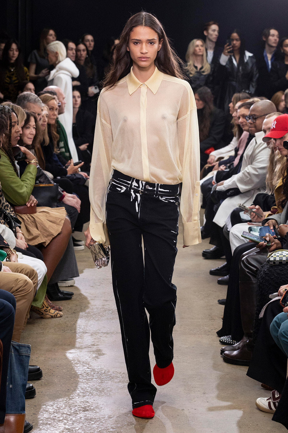 Female runway model walking a fashion show in a sheer beige blouse, black paint-splattered pants, and bright red shoes, with seated audience on both sides.