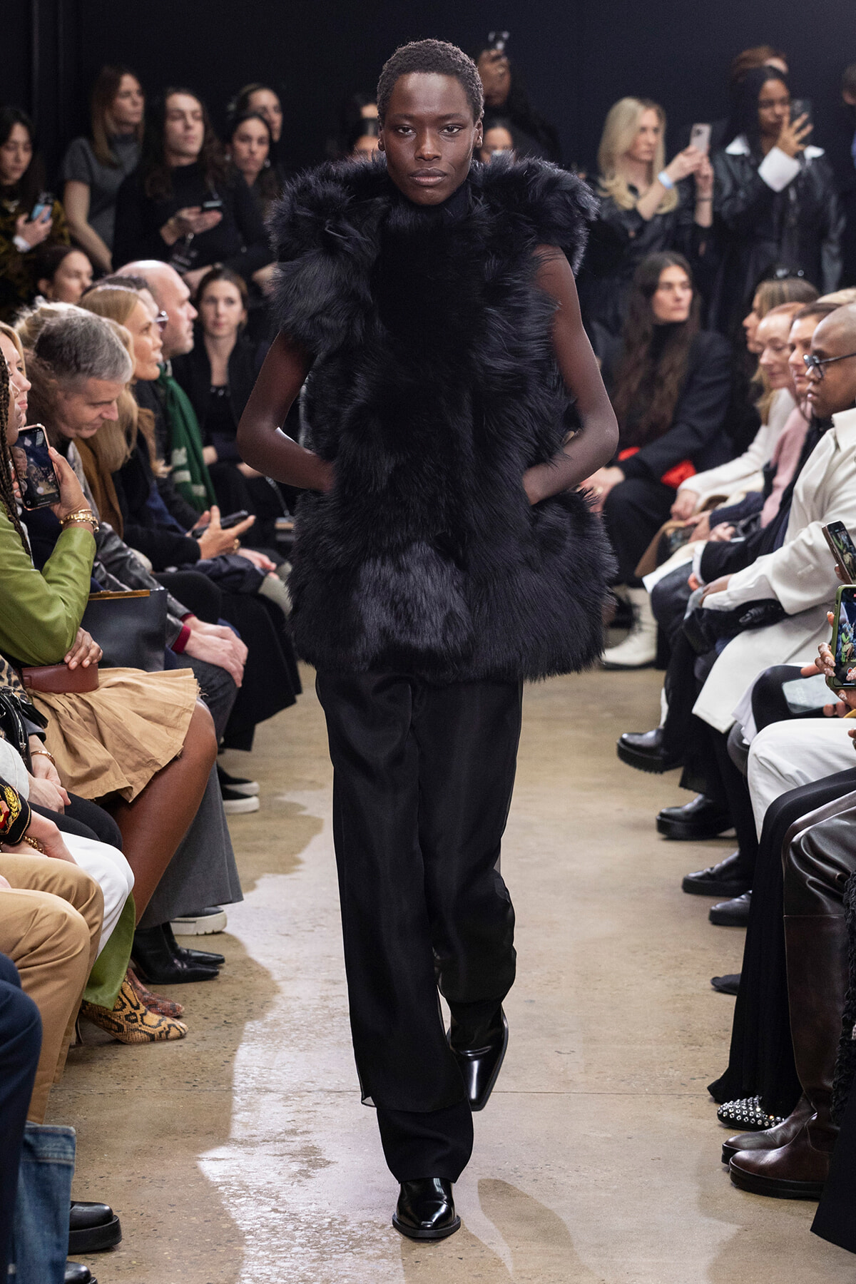 Model walks the runway in a dark fur vest and black trousers while audience members snap photos and watch.