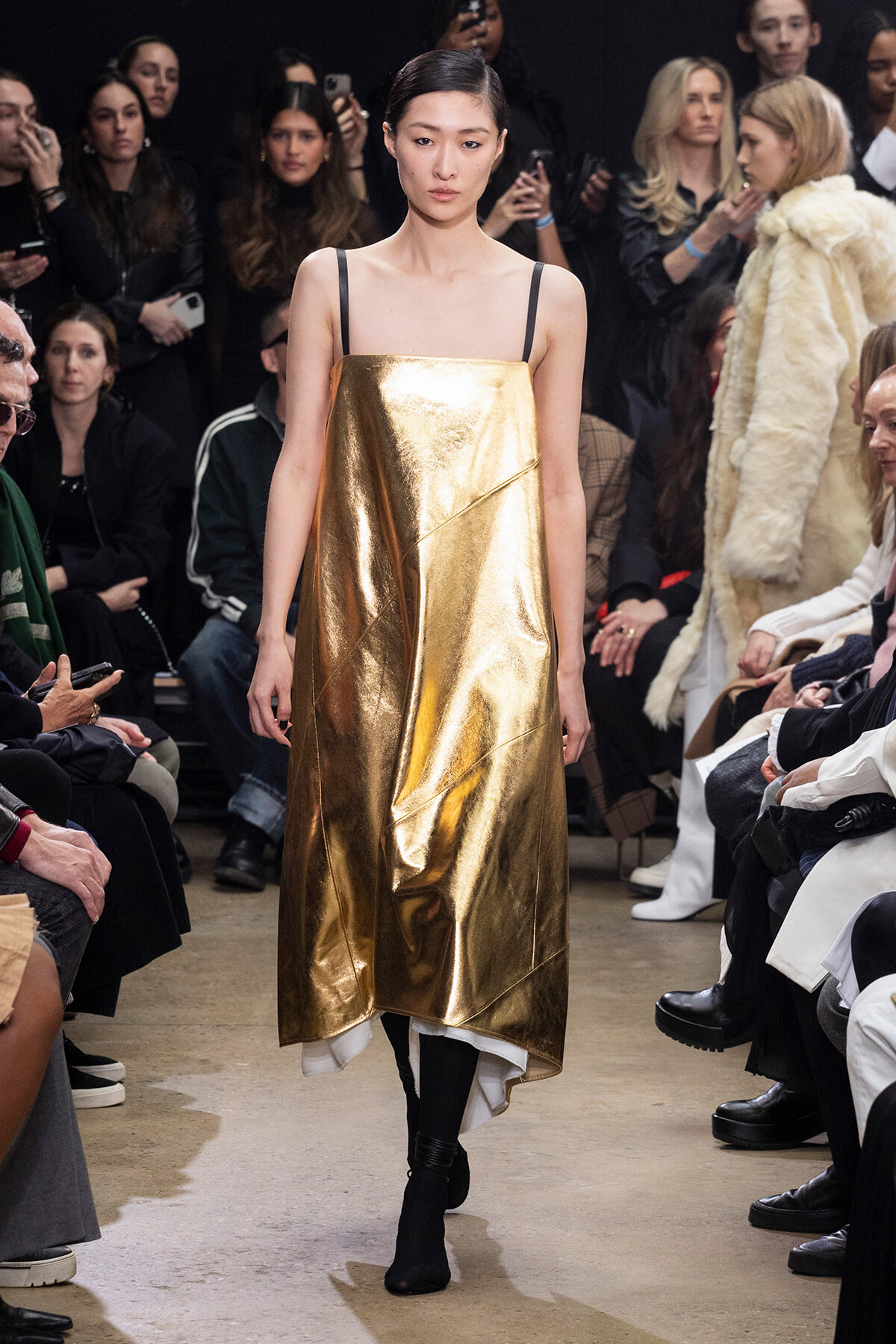 Model walks the runway wearing a shiny gold metallic dress with thin black straps, surrounded by an audience.