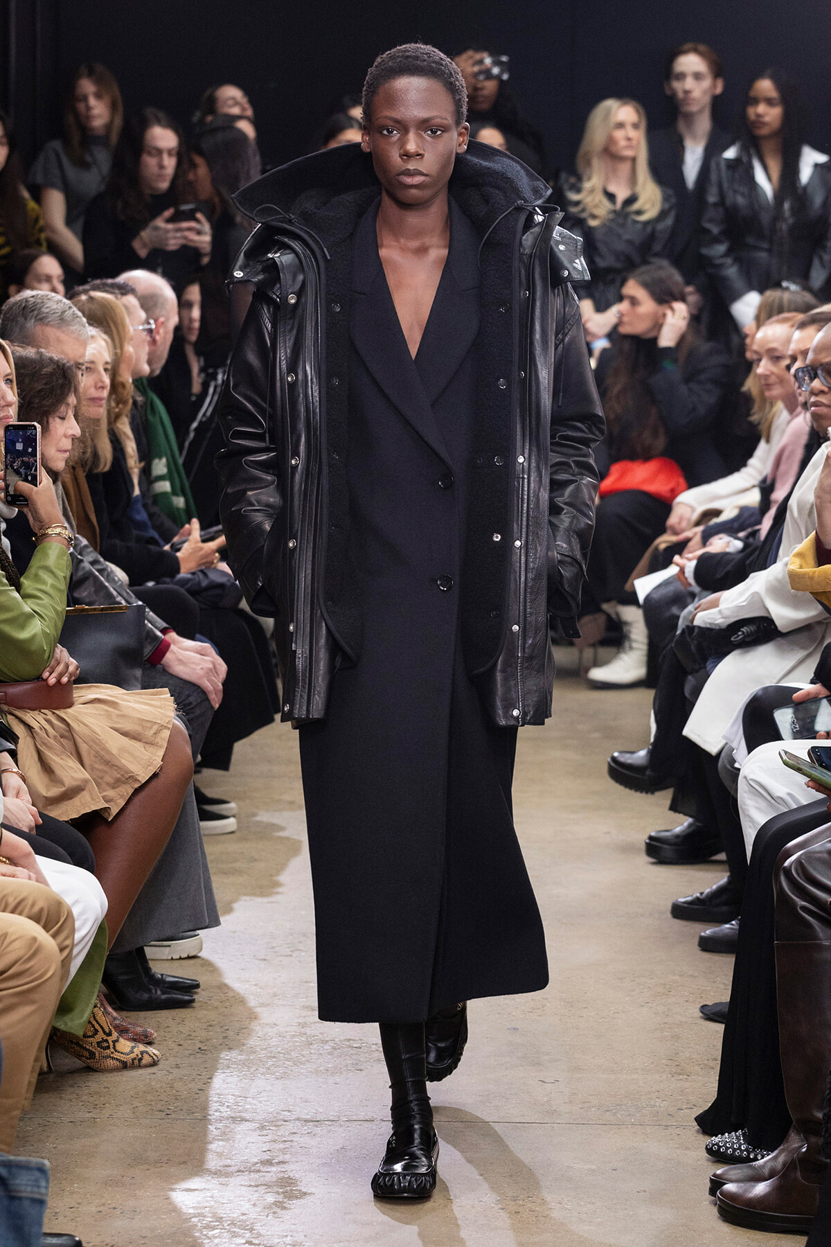 Model walking a runway in a long black outfit with an oversized black leather jacket, surrounded by seated audience on both sides of the catwalk.