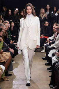 Model walks runway in an all-white suit with a white turtleneck, audience seated on both sides of the catwalk