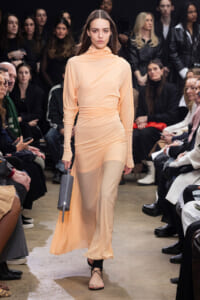 Model walks a peach satin long-sleeve gown down the runway, holding a gray cylindrical clutch as seated audience watches on both sides.