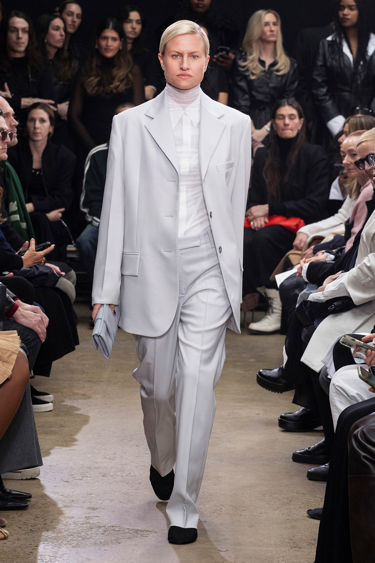 Model walks a runway in a light gray, oversized suit with a white turtleneck, holding a matching clutch.