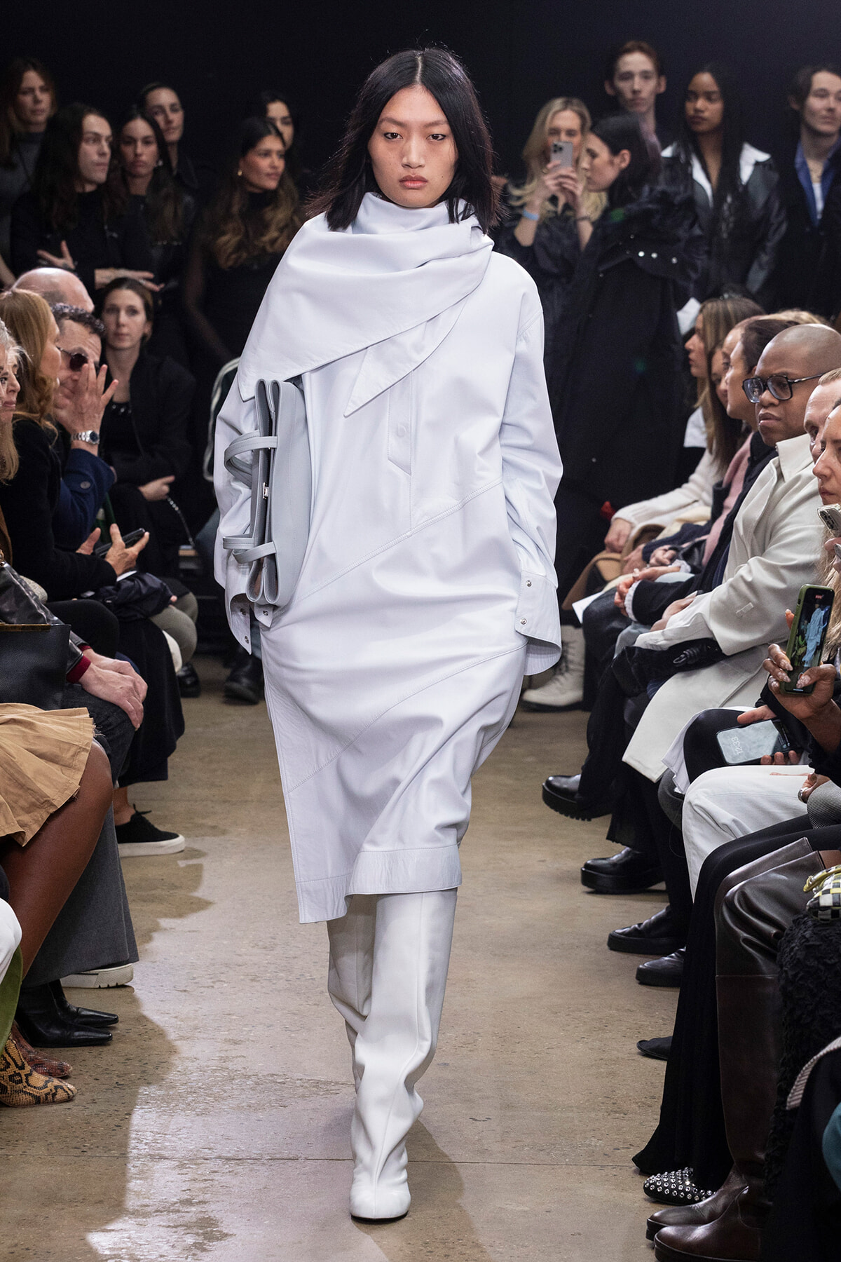 Model walking the runway in a white, oversized coat and matching trousers, audience seated on both sides watching the show.
