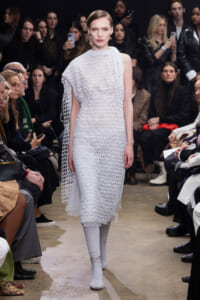 Model walks the runway in a white crochet dress with an attached cape during a fashion show, with an audience watching on both sides.
