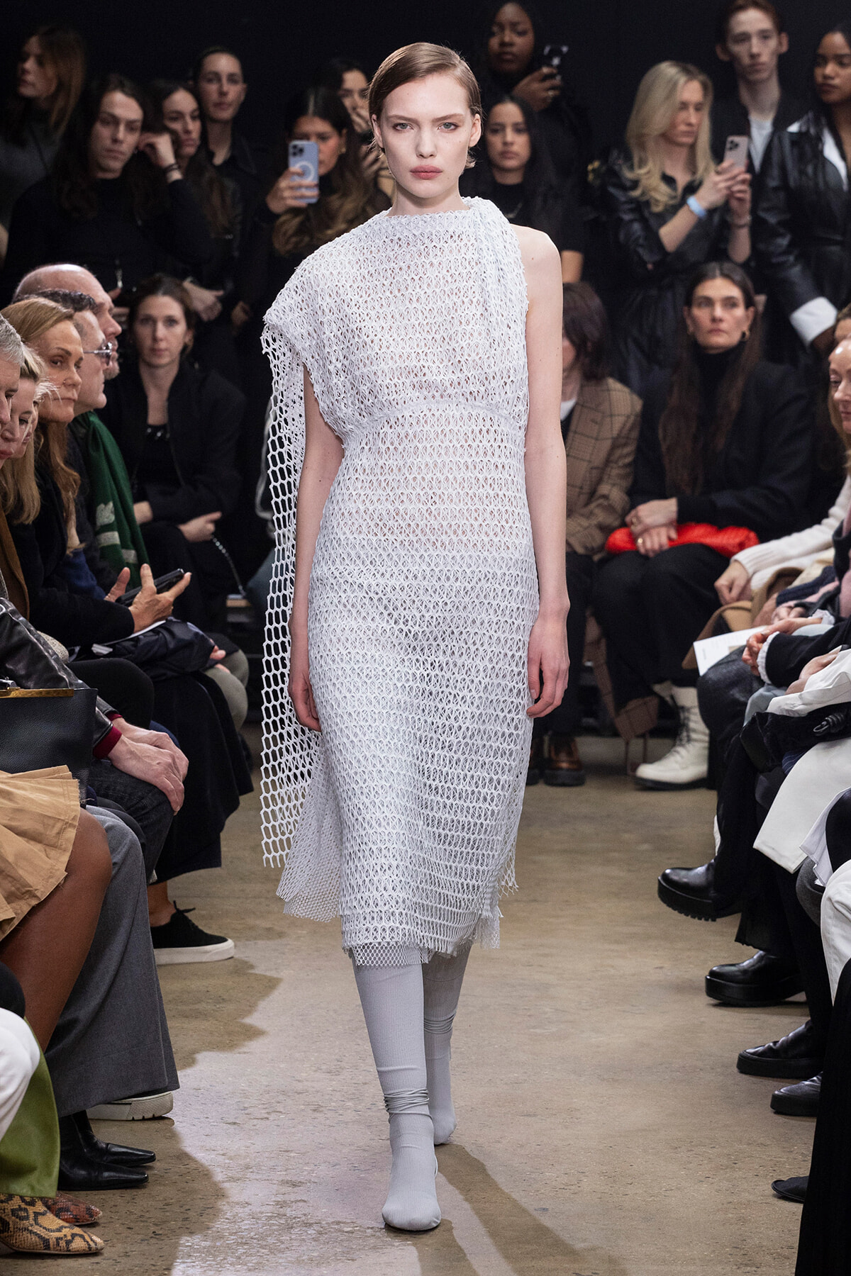 Model walks the runway in a white crochet dress with an attached cape during a fashion show, with an audience watching on both sides.