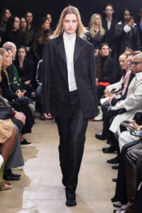 Female model walks a runway in an oversized black blazer and white shirt, with textured black pants as spectators watch from the sides.