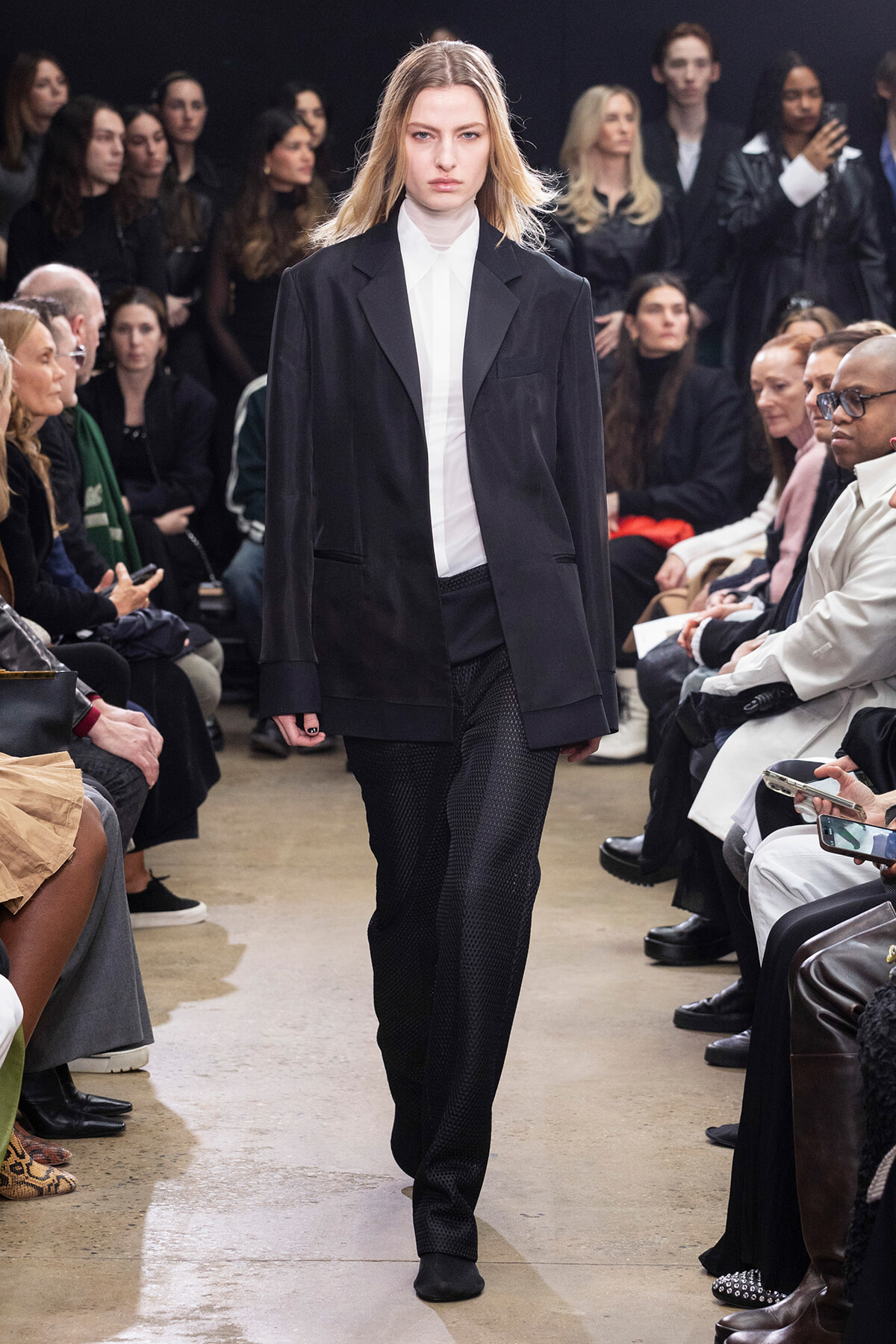 Female model walks a runway in an oversized black blazer and white shirt, with textured black pants as spectators watch from the sides.