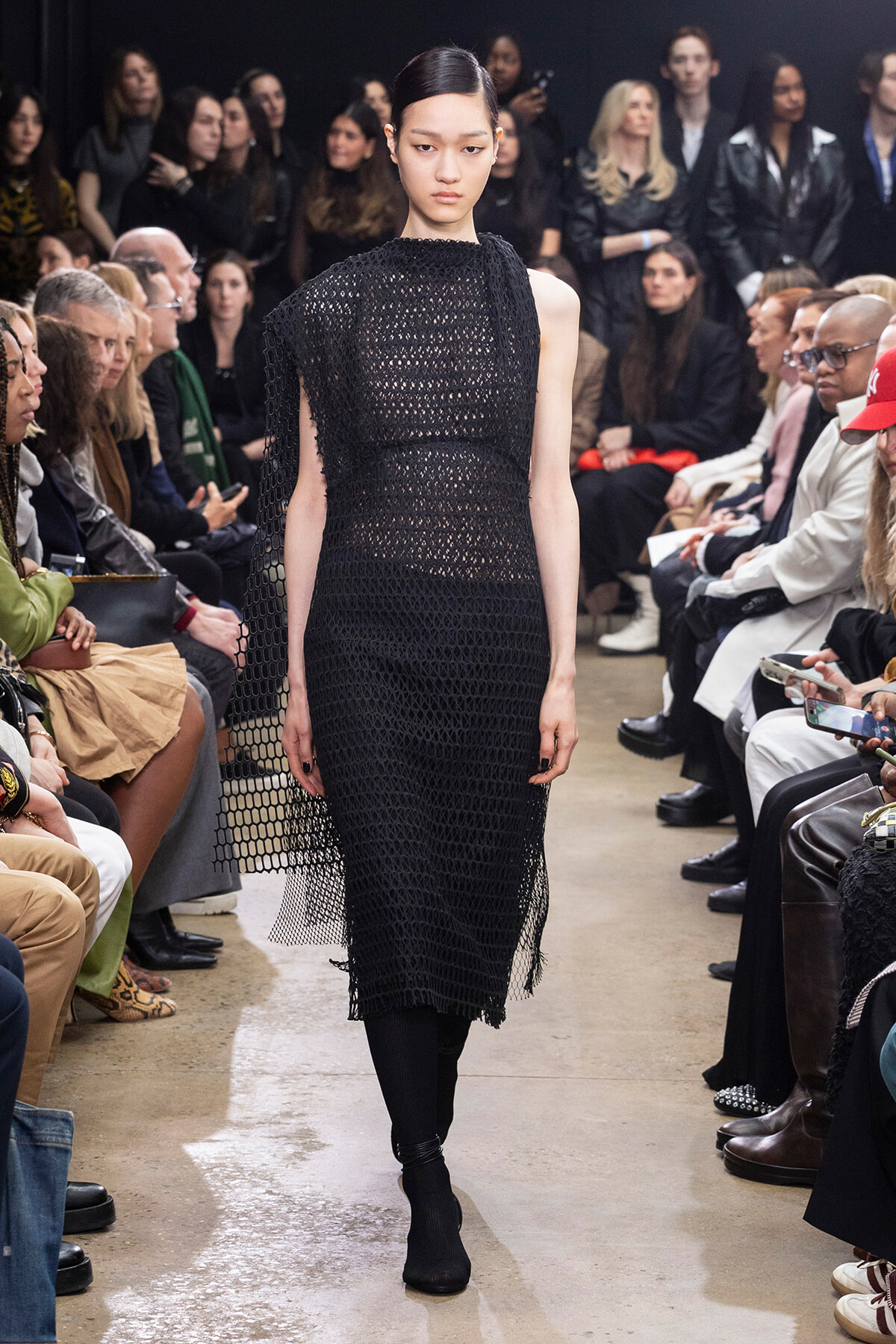 Model walks a fashion runway in a black open-weave mesh dress with spectators on both sides.