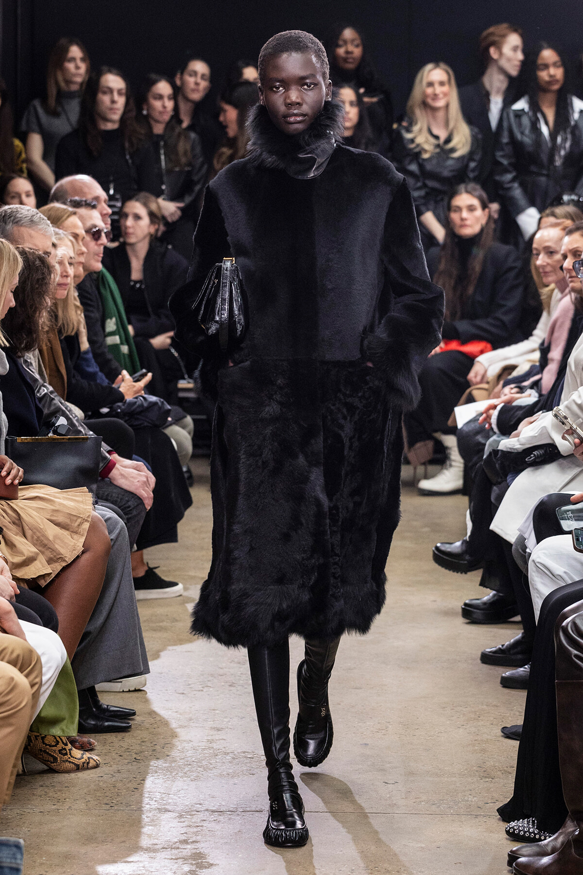 Model walks the runway in a long black fur coat with a high collar, as seated audience members watch on both sides.